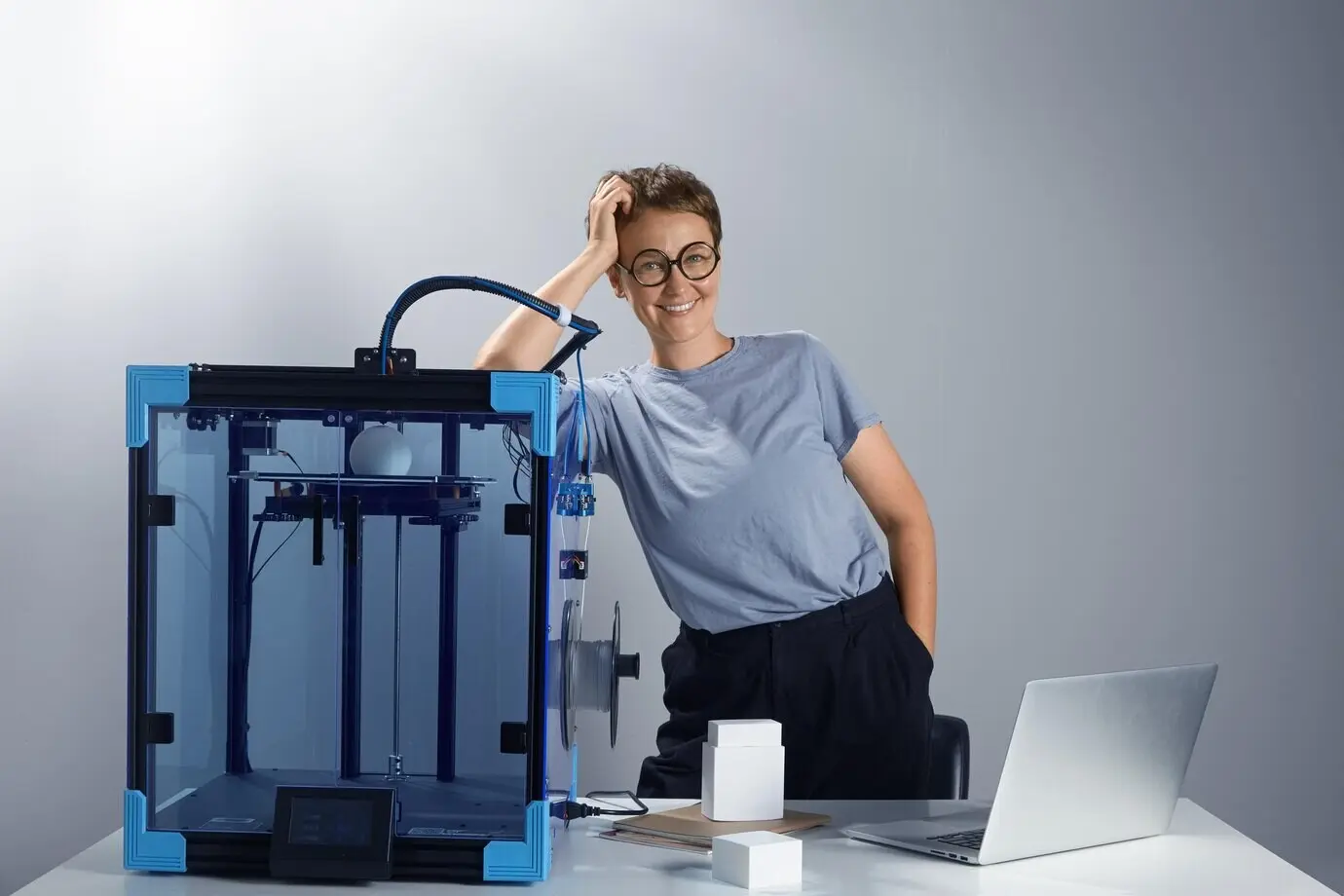 Schöne kaukasische Frau mit Brille lächelt neben einer 3D-Druckmaschine in ihrem Büro; Konzept neuer Technologien für kleine Unternehmen; Computer-Prototypen auf dem Tisch; technisches Ambiente; JPG-Foto.